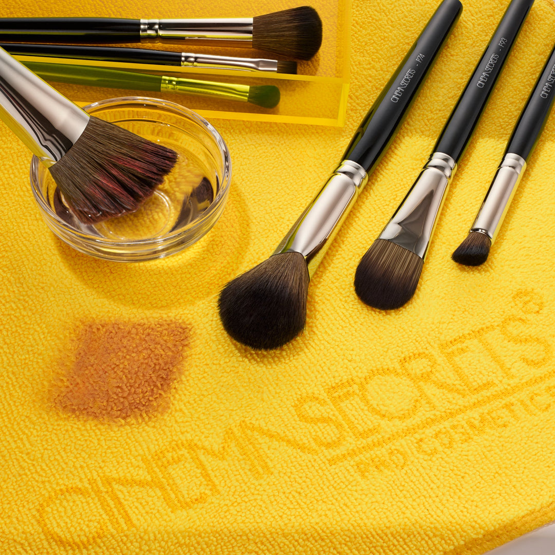 cinema secrets makeup brush cleaner being used to clean brushes. White background, with one hand holding a brush, dipping it in a glass bowl with 4 makeup brushes surrounding.