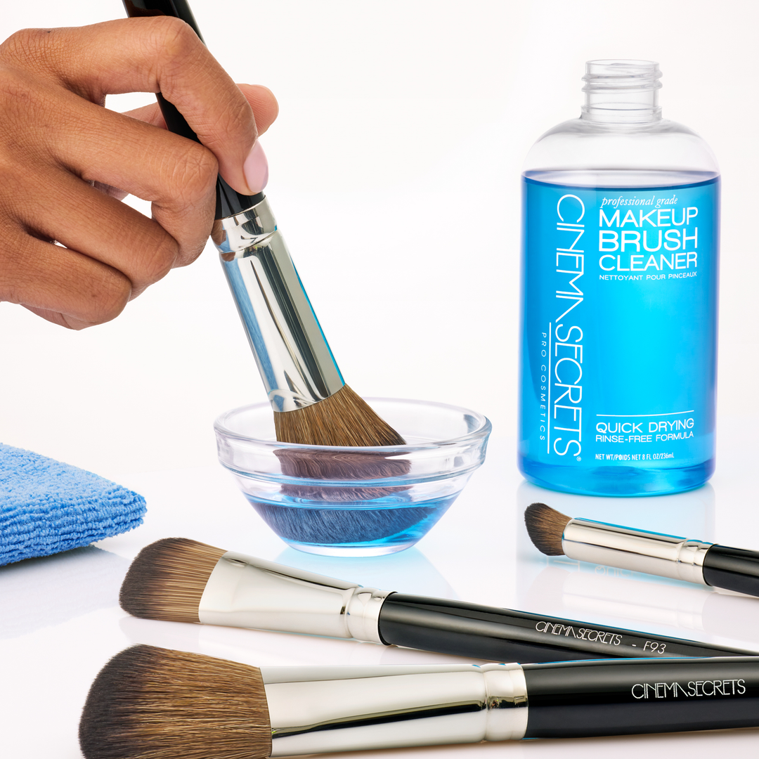 cinema secrets makeup brush cleaner being used to clean brushes. White background, with one hand holding a brush, dipping it in a glass bowl with 4 makeup brushes surrounding.